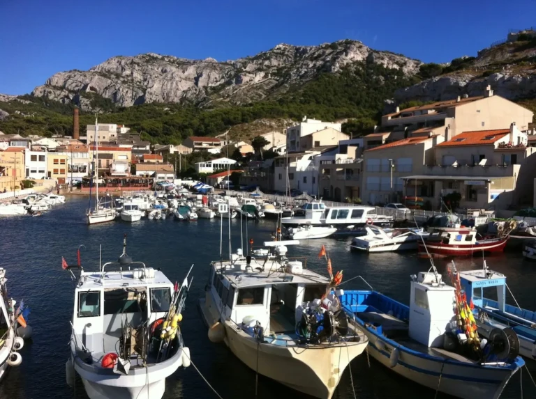 Vue du port de la Madrague de Montredon à Marseille, avec des bateaux de pêche amarrés et des maisons typiques en arrière-plan.