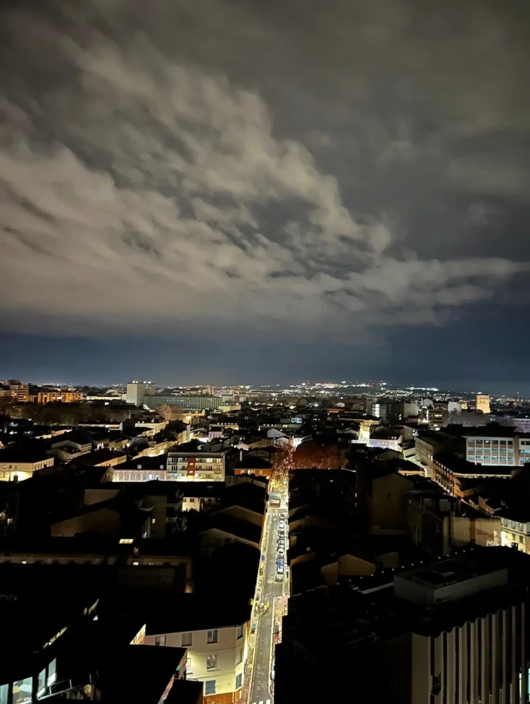 Vue nocturne sur les toits de Toulouse depuis un penthouse, avec nuages et éclairage urbain.